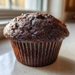 A single, dark, moist chocolate muffin topped with chocolate chips, sitting on a counter near a window.