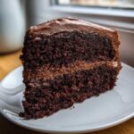 A close-up of a rich, moist slice of homemade chocolate cake layered with chocolate frosting.