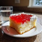 A moist slice of jello poke cake topped with bright red gelatin and white whipped cream, served on a white plate.
