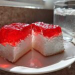 Two square slices of jello poke cake featuring a white creamy layer and a bright red, glossy jello topping, served on a white plate.