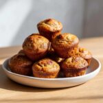 A pile of golden-brown, moist mini banana muffins stacked high on a light-colored plate.