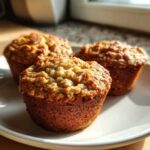Close-up of three golden brown, moist oatmeal muffins with textured oat topping, sitting on a white plate near a window.