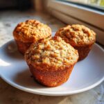 Three golden brown, moist oatmeal muffins with visible oat flakes on top, sitting on a white plate near a window.
