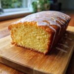 A close-up of a moist, golden olive oil cake loaf, drizzled with white lemon glaze, resting on a wooden cutting board.