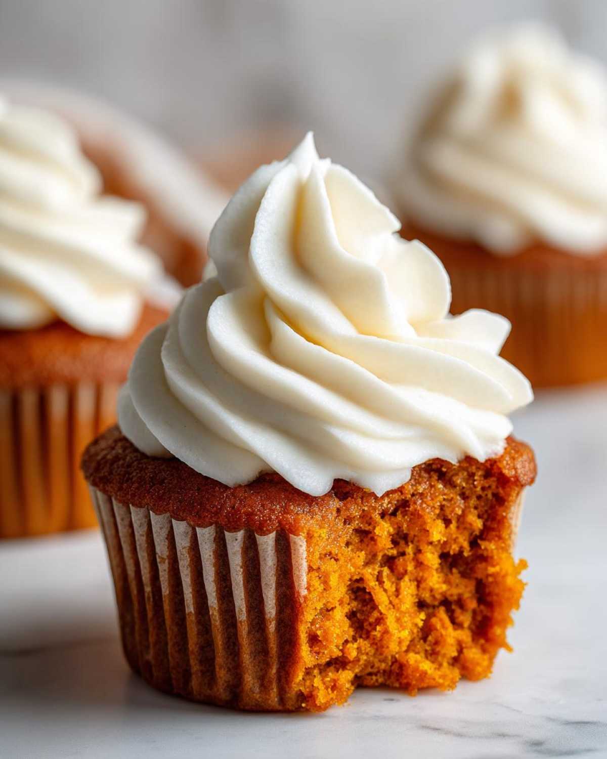 A close-up shot of a moist pumpkin spice cupcakes with a bite taken out, topped with thick white cream cheese frosting.