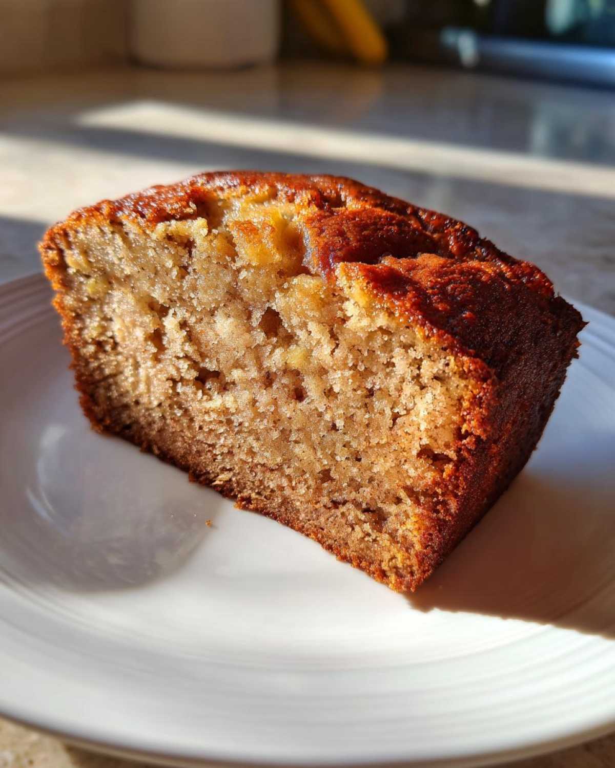 A close-up slice of moist, golden-brown banana bread recipe on a white plate, highlighted by sunlight.