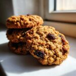 A close-up stack of three textured oatmeal raisin cookies resting on a white surface near a window.