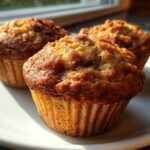 Close-up of three freshly baked, golden brown apple muffins sitting on a white plate near a window.