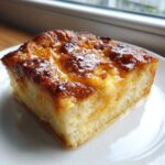 A close-up of a square slice of baked overnight french toast, topped with a shiny, caramelized glaze.