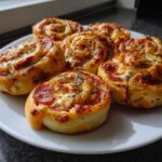Close-up of several freshly baked pepperoni pizza pinwheels topped with melted cheese and herbs on a white plate.