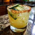 Close-up of a frosty Pickle margarita in a rocks glass with a chili-salt rim and cucumber slices.