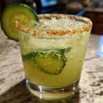 Close-up of a bright green Pickle Margarita in a glass with a chili-salt rim and cucumber slices.