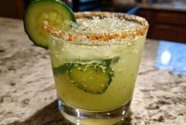 Close-up of a bright green Pickle Margarita in a glass with a chili-salt rim and cucumber slices.
