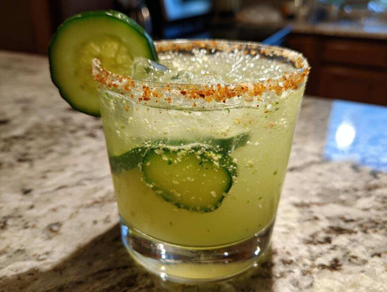 Close-up of a bright green Pickle Margarita in a glass with a chili-salt rim and cucumber slices.