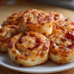 A close-up of several freshly baked, golden brown pizza pinwheels piled on a white plate.