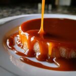 Thick, glossy caramel sauce being poured over a small cake or flan, creating drips down the side.