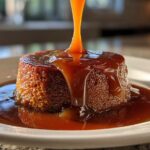 A stream of rich caramel sauce is being poured over a small, round cake or pudding, creating a glossy pool.
