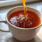 Close-up of rich, dark amber bone broth recipe being poured into a white mug, showing its thick, gelatinous consistency.