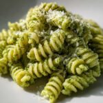 Close-up of fusilli protein pasta tossed in bright green pesto and topped with grated Parmesan cheese.