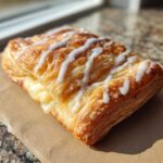 Close-up of a golden, flaky puff pastry danish with cream cheese filling and white icing drizzle.