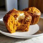 A close-up of a Pumpkin Cream Cheese Muffin cut in half showing the creamy filling, with another muffin behind it.
