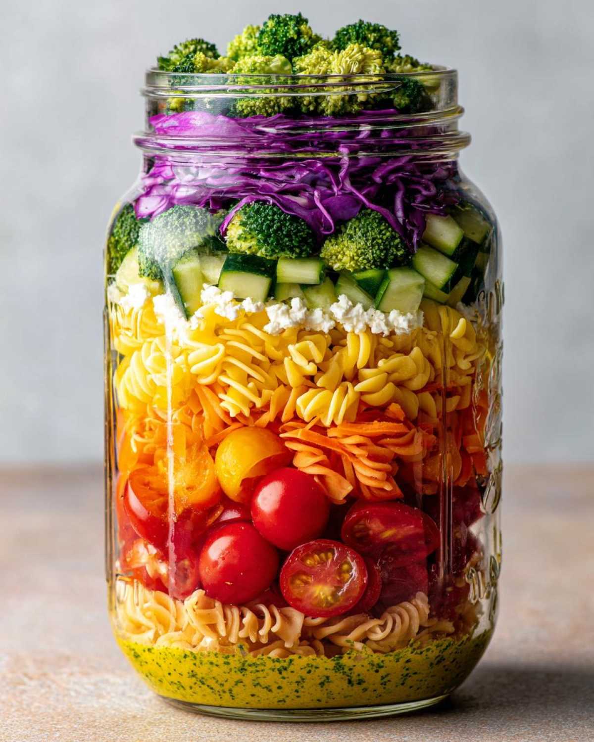 A tall glass jar layered with colorful ingredients for a Rainbow Veggie Pasta Salad, showing dressing, pasta, tomatoes, carrots, cheese, cucumber, cabbage, and broccoli.