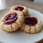 Three buttery raspberry thumbprint cookies with bright red jam centers resting on a simple white plate.