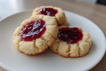 Three buttery raspberry thumbprint cookies with bright red jam centers resting on a simple white plate.
