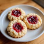 Close-up of three delicious raspberry thumbprint cookies with bright red jam centers on a light gray plate.