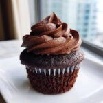 A single, moist chocolate cupcake topped with a generous swirl of dark chocolate frosting, sitting on a white plate.