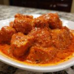 Close-up view of tender chicken pieces coated in a vibrant, orange-red, creamy sauce, characteristic of authentic butter chicken.
