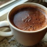 Close-up of a creamy, rich hot chocolate with foam bubbles in a light-colored mug by a window.