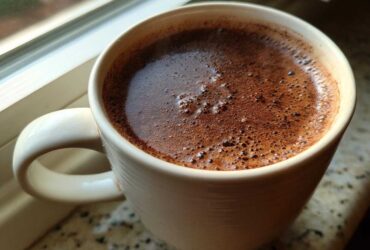Close-up of a creamy, rich hot chocolate with foam bubbles in a light-colored mug by a window.