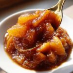 Close-up of thick, dark brown apple pie filling being lifted out of a white bowl on a golden spoon.
