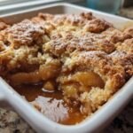 Close-up of a ridiculously delicious apple cobbler with a crumbly topping and gooey filling in a white baking dish.