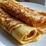 Close-up of three golden, rolled cottage cheese wrap crepes stacked on a white plate.