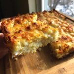 A hand holds up a square slice of cheesy, baked hashbrown casserole showing its creamy interior and golden-brown top.