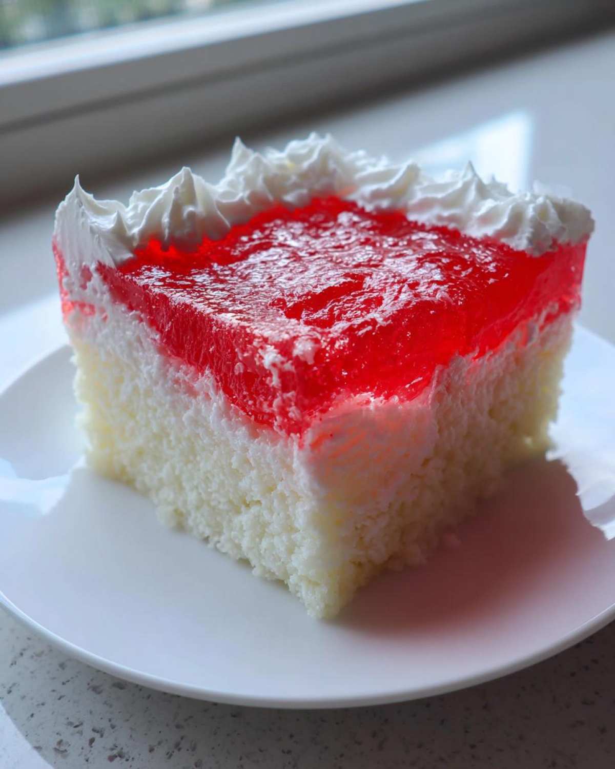 A square slice of moist jello poke cake featuring a white cake base, a layer of red gelatin, and piped whipped cream on top.