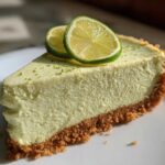 A close-up of a perfect slice of creamy key lime cheesecake on a white plate, topped with lime zest and two thin lime slices.