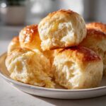 Close-up of golden brown, soft buttery rolls piled on a white plate, one roll is pulled apart showing the fluffy interior.