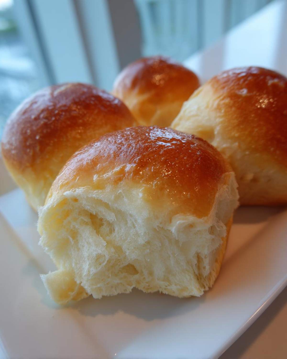 Close-up of four soft, buttery rolls, one pulled apart to show the fluffy interior and shiny glaze.