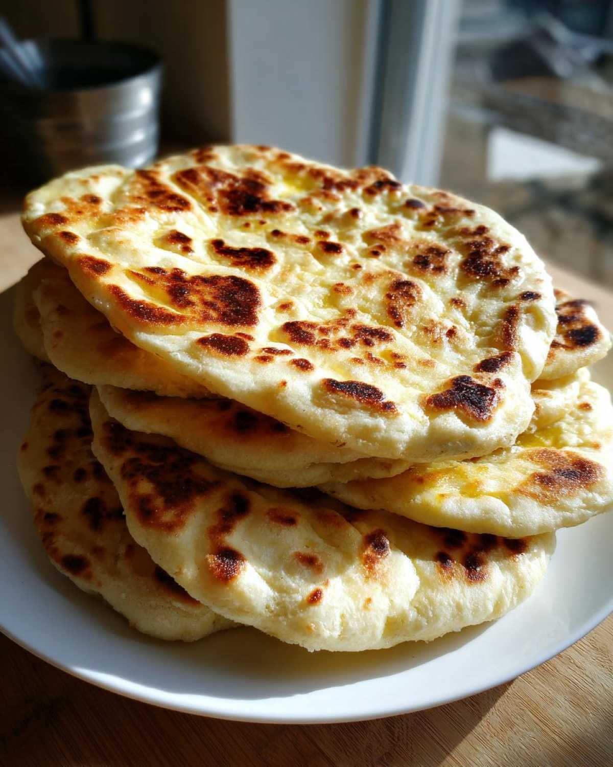 A stack of freshly made, soft naan bread with beautiful golden-brown charred spots, resting on a white plate.