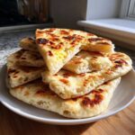 A stack of freshly baked, soft naan bread pieces piled on a white plate, showing charred spots.