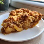A close-up of a warm slice of Spiced Pear Crumble on a white plate, showing soft spiced pears and a thick, crunchy topping.