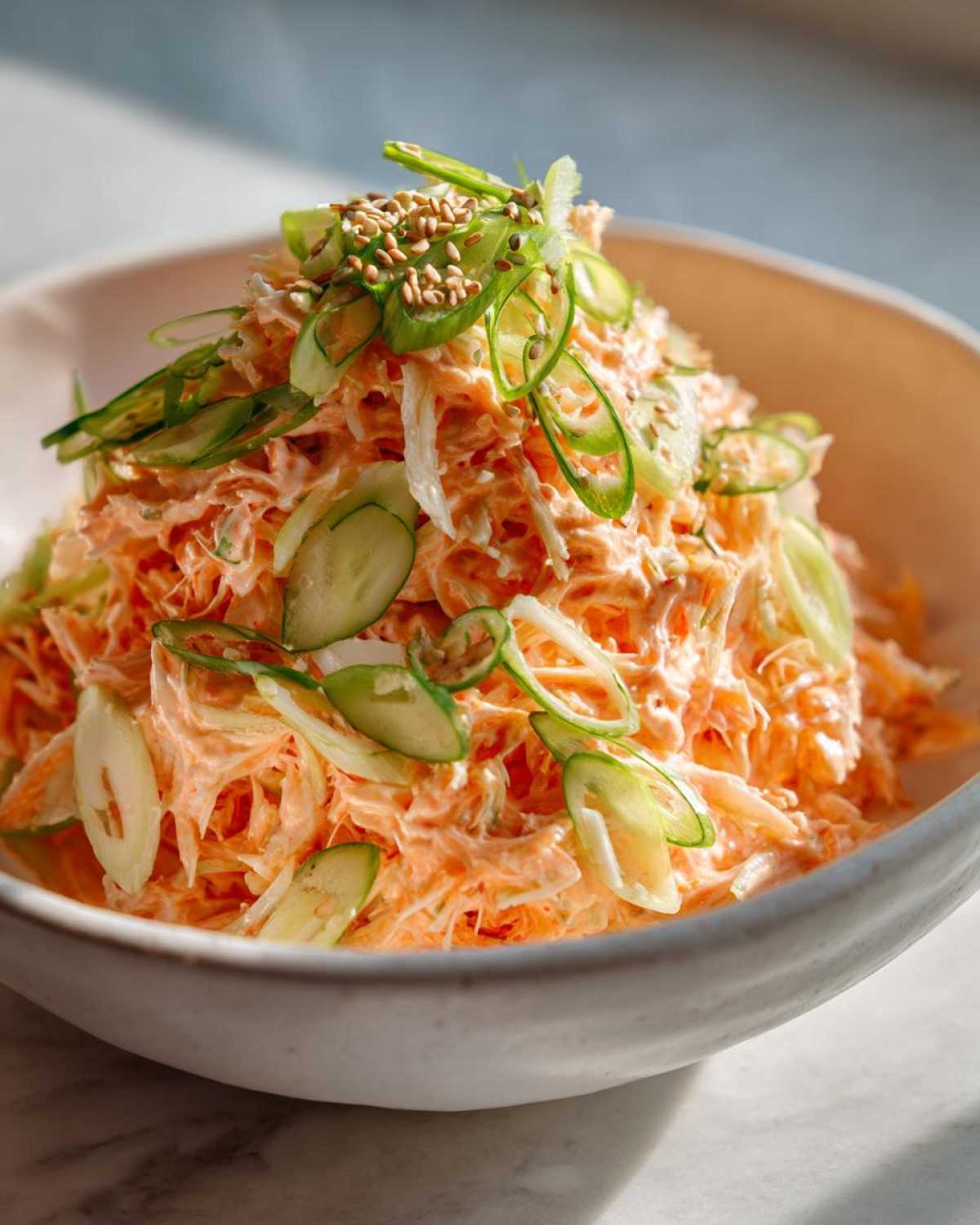 A mound of creamy, orange-tinted spicy kani salad garnished with sliced cucumbers, green onions, and toasted sesame seeds.