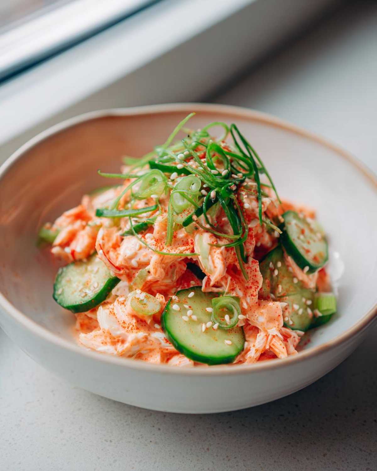 A close-up of spicy kani salad mixed with creamy dressing, sliced cucumbers, and topped with green onions and sesame seeds.