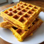 Close-up of a stack of three golden, fluffy pumpkin waffles resting on a white plate.