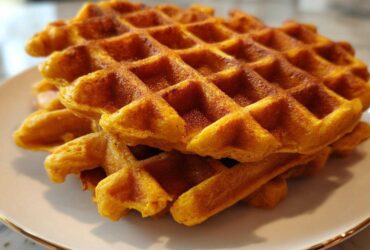 A close-up stack of golden brown, perfectly textured pumpkin waffles on a white plate.
