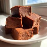 A stack of rich, brown squares of homemade penuche fudge piled on a white plate, highlighted by natural sunlight.