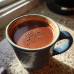 Close-up of rich, steaming hot chocolate with foam in a textured blue mug, illuminated by sunlight.